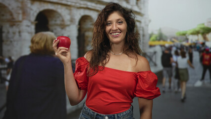 Woman smiling holding red apple and pointing finger at coliseum building on a busy tourist street;...