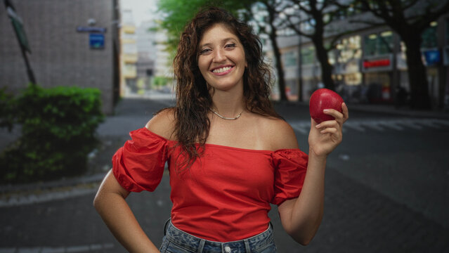 Woman holding red apple on a city street wearing denim and a necklace, smiling with exposed shoulders and hands visible; happiness.