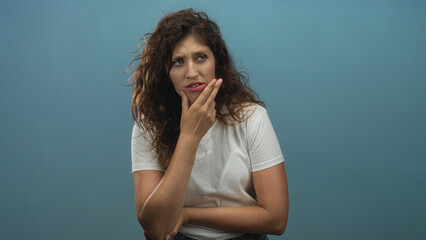Woman touching chin with fingertips and crossed arms in studio against teal blue backdrop; thoughtful concern.