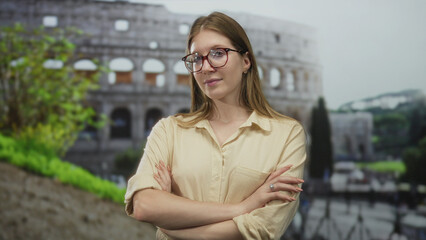 Woman in beige shirt and glasses stands with arms crossed before coliseum building outdoors in...