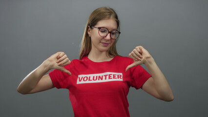 Woman in red volunteer shirt standing confidently with thumbs pointing to chest in a grey studio; pride.