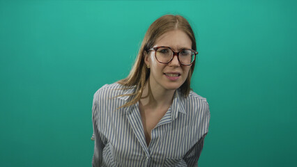 Blonde woman tilts head and brushes hair while adjusting glasses against teal studio backdrop;...