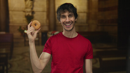 Man in red shirt holds a glazed donut with his right hand while smiling beside a wooden pew in an old church; joyful devotion.
