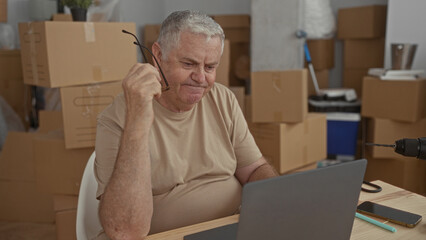 Man holding reading glasses near face while studying laptop at a cluttered table surrounded by packed moving boxes in a building; frustration.