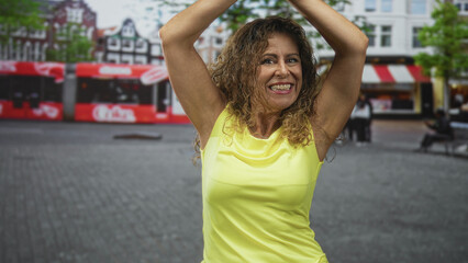 Woman with raised arms showing armpit and smiling in street while wearing yellow tank top; joy...