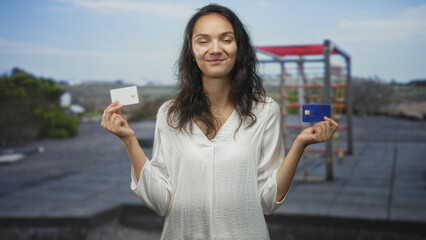 Woman holding a white card and a blue creditcard in each hand, looking contemplative on a building rooftop; decision confidence.