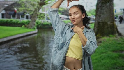 Young woman in yellow crop top and denim overshirt frames with hands and smiles showing midriff on street by canal and tree lined path; playful confidence.
