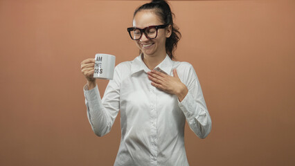Woman in white shirt holding a mug that reads i am the boss with hand on chest gesture and wide smile in studio; confidence leadership.