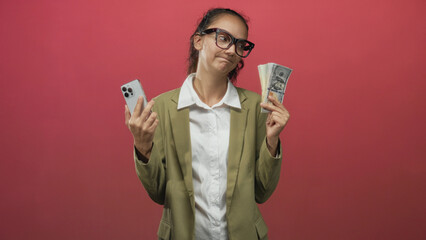 Woman holding smartphone and a fan of cash, shrugging with raised shoulders in studio; indecision materialism choice.