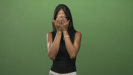 Thai woman with glasses pressing hands to mouth in studio green screen, bare forearms visible and black sleeveless tanktop; anxiety vulnerability.