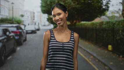 Thai woman smiling with bare shoulders and head tilted, striped tank top visible, standing on a city street lined with parked cars; calm confidence.