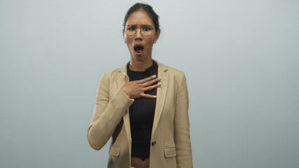 Thai woman hand on chest with open mouth and glasses wearing beige blazer in studio; surprise...