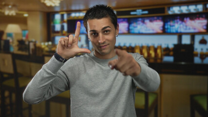 Young hispanic man frames camera lens with l hand gesture and points finger to camera in...