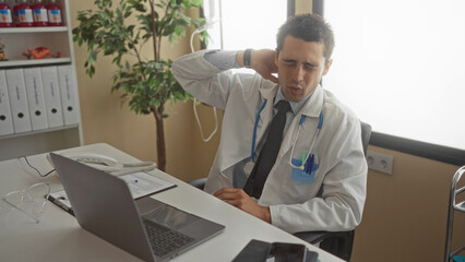 Young man doctor in uniform with stethoscope in clinic room appears stressed holding neck near...
