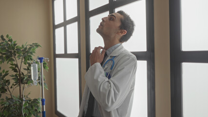 Young hispanic man doctor in white coat with stethoscope removes glasses in a clinic building near an iv stand by a window; fatigue.