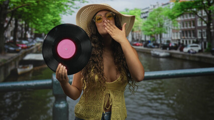 Hispanic woman holding vinyl record with hand, smiling and waving on a street bridge while wearing sunhat and sunglasses; joyful carefree summer.