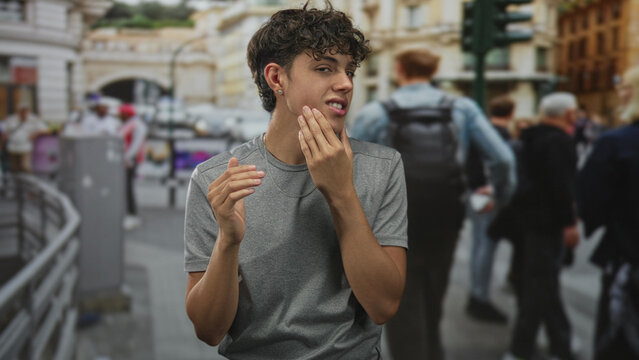 Teen boy touching cheek with hand, grimacing while standing on a busy street with passersby; awkward introspection.