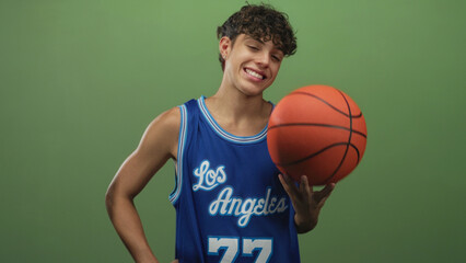 Naklejka premium Teenage boy balancing an orange basketball on his fingers while smiling, wearing a blue los angeles jersey against a green studio backdrop; playful confidence.