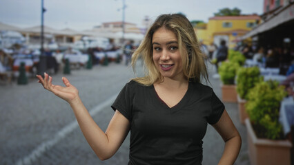 Fototapeta premium Young blonde woman points finger and raises open hand on a cobblestone street market while smiling and engaging with camera; friendly welcome.