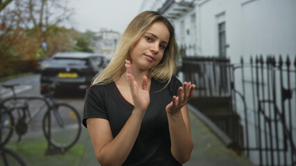 Blonde woman clapping hands on a narrow city street near bicycles, iron railings and a parked car, wearing black t shirt; urban contentment.