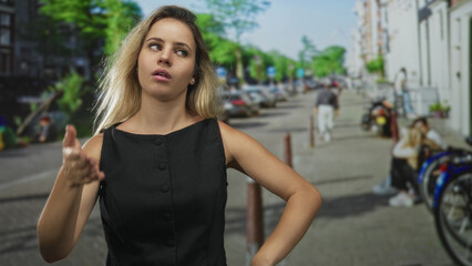 Woman young blonde adjusts dress collar with both hands while standing near parked bicycles on a busy city street; defiance attitude.