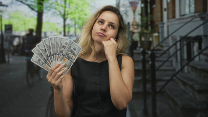 Young blonde woman holding us dollar bills with hand on chin in busy city street; pensive wealth...