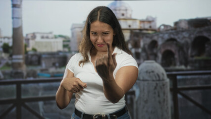 Fototapeta premium Young latina woman shows middle finger and points with other hand at ancient ruins building on outdoor terrace while smiling; defiance.