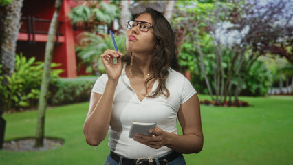 Fototapeta premium Woman wearing glasses holding pen to temple and reading a notepad in forest near a red building and palm trees; pensive concentration.