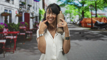 Senior hispanic woman smiling, holding a pottery bottle to her temple and pointing index finger to her cheek in a street cafe with tables, chairs and trees, wearing a white blouse and bracelets; joy.