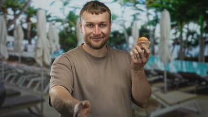 Man holding muffin with outstretched hand in resort building poolside among loungers and umbrellas by the turquoise water; relaxation holiday.