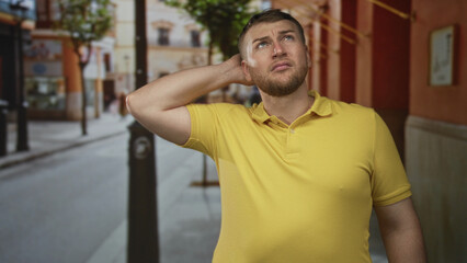 Fototapeta premium Man touching cheek with hand while looking up in city street by lamppost and storefront, wearing yellow polo shirt; concern.