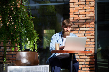 Man works on laptop at outdoor round table, with glass of iced coffee. Solar panel nearby, emphasizing sustainable, modern eco-friendly workspace that combines technology and sustainability.