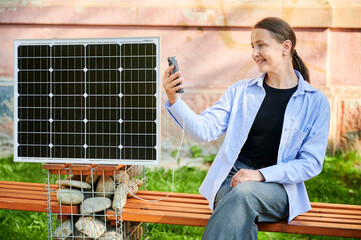 Happy woman using smartphone connected to photovoltaic solar panel. Integration of sustainable renewable energy into everyday life, demonstrating practical use of solar power for charging devices.