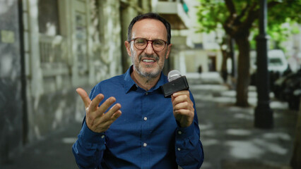 Hispanic man in blue shirt speaking passionately into microphone on a sunny urban street with blurred city background.