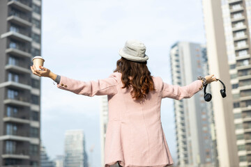 back view of a woman in a pink coat with her arms spread wide while holding headphones in one hand and a coffee in the other in a modern city setting with modern skyscrapers. 