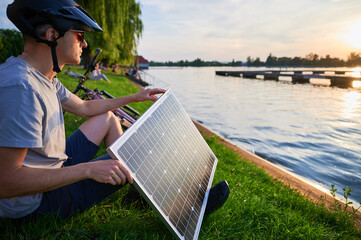 Man cyclist using solar panel for charging electric mountain bike outdoors. Guy in helmet and sunglasses sits by lakeside, enjoying tranquil scene as sun sets over water.