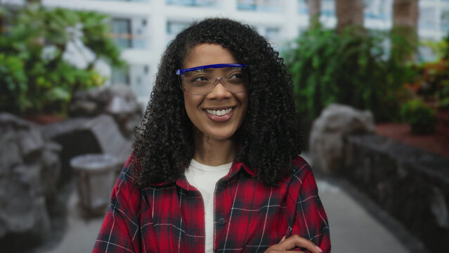 Woman wearing safety glasses smiles and crosses arms in a sunlit green outdoor park setting; confidence. - Powered by Adobe