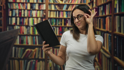 Young woman holding black book and adjusting glasses while smiling among tall bookshelves in a library building  curiosity. © Krakenimages.com