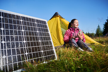 Happy woman sits in front of yellow tent, charging her phone by using solar panel, highlighting use of renewable energy and eco-friendly camping. On background lush green trees and clear blue sky.