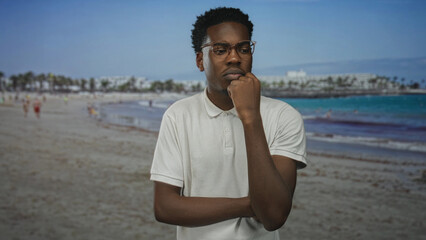 Young man wearing white polo shirt with hand on chin gesture at sunlit coastal building; self reflection calm.