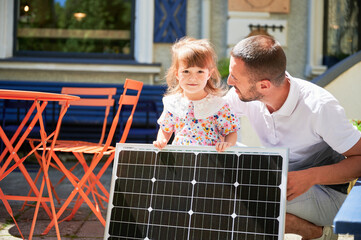 Young father showing his kid solar panel. Dad smiling with his cute daughter next to portable solar battery. Concept of sustainable lifestyle and green renewable energy.