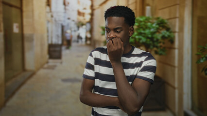 Man in striped shirt bites fingernails on narrow city street lined with stone walls under warm...