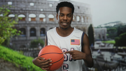 Young african american man wearing usa jersey points finger to basketball beside building in front of roman coliseum; pride.
