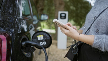 Woman using smartphone at electric car charging station in outdoor parking with black vehicle showing plugged charger cable.