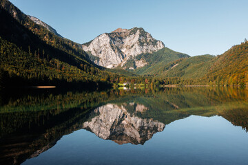 Peaceful alpine lake reflecting the rugged Höllengebirge mountains, surrounded by dense green forest and a small lakeside house under clear blue sky. High quality stock photo