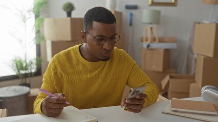 Man writing notes with smartphone in studio stacked with moving boxes and notebooks; concentration productivity.
