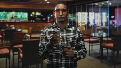 Young black man holds smartphone and takeaway coffee, fingers on screen and hand visible while standing in a building interior; concentration and routine.