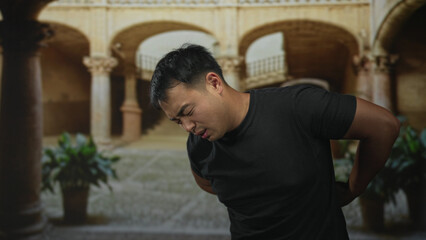Young chinese man in black shirt winces and holds lower back amid weathered stone arches in...