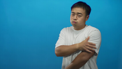 Young chinese man holds elbow in bright blue studio setting while pressing his forearm with his other hand for support and subtle tension in posture; serenity.