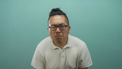 Fototapeta premium Man with frowning face and squinted eyes in a teal studio backdrop, wearing glasses and white polo shirt, seated and leaning slightly forward; frustration.